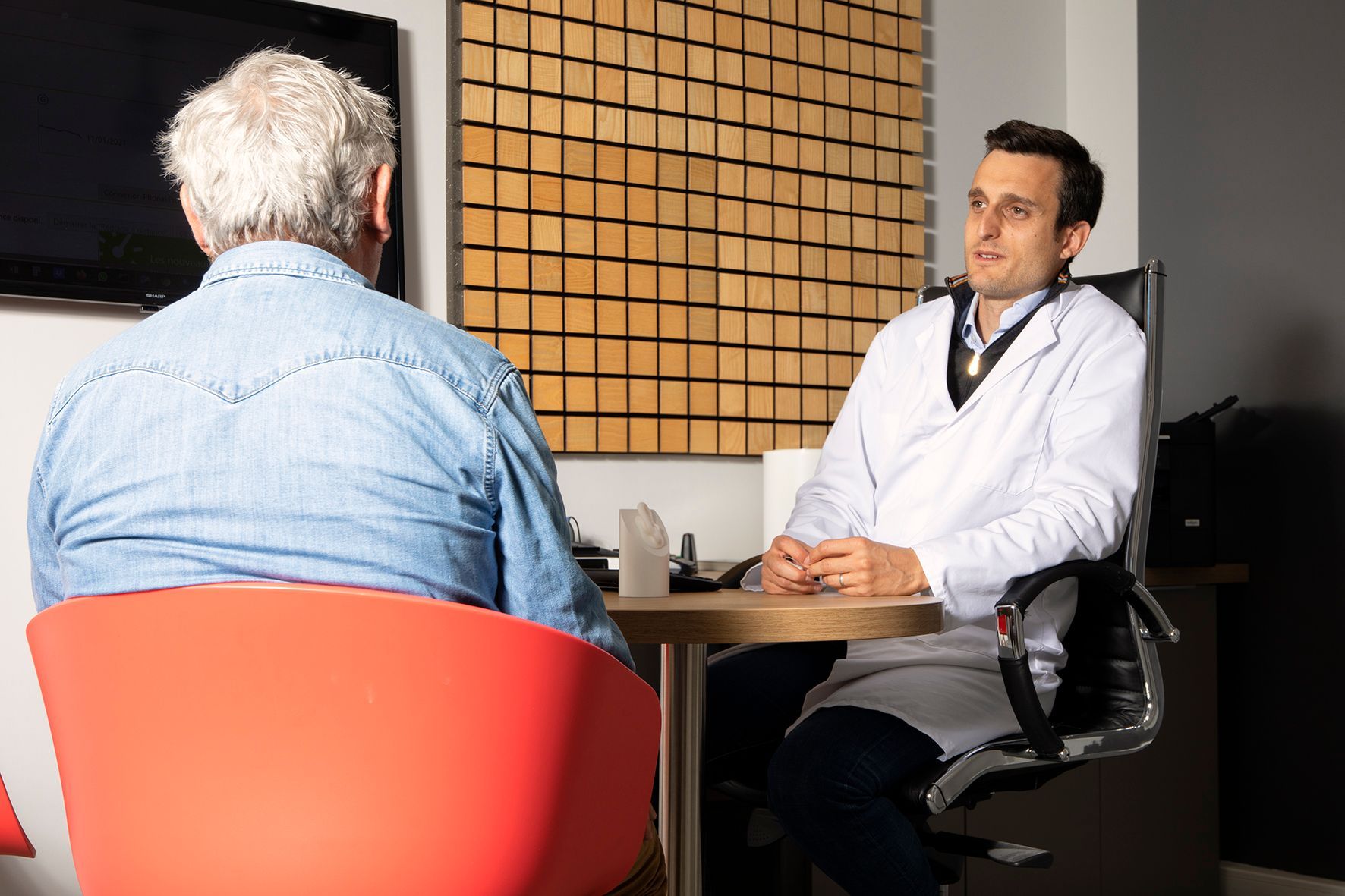 Un médecin parle à un patient assis dans son bureau, un ordinateur portable sur le bureau et un mur de notes derrière lui.