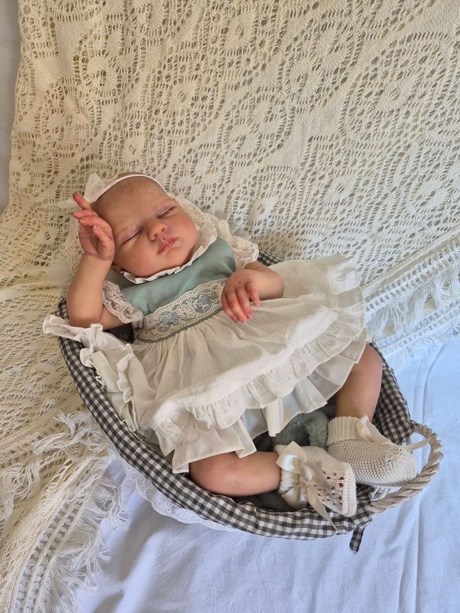 Muñeca bebé durmiendo con los ojos cerrados, una manta de punto azul y una mano levantada sobre una superficie blanca y esponjosa.