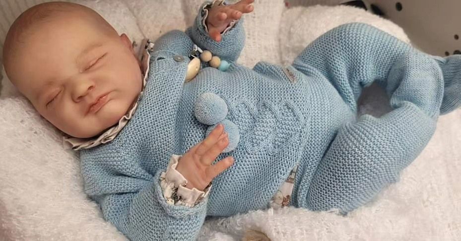 Muñeca bebé durmiendo con un traje de punto azul sobre una manta blanca.