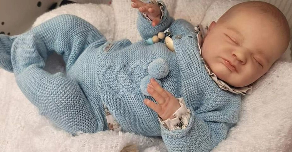 Bebé durmiendo con un conjunto de punto azul, tumbado sobre una manta blanca, sosteniendo un pequeño peluche.