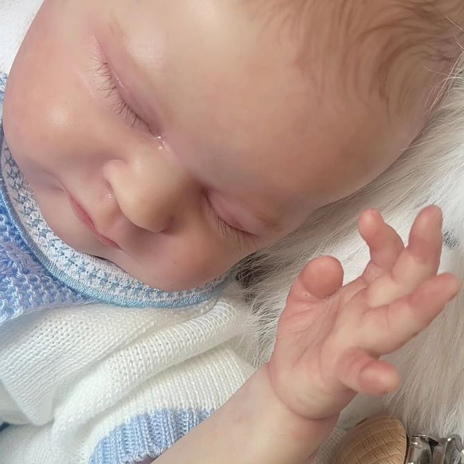 Muñeca bebé durmiendo con los ojos cerrados, una manta de punto azul y una mano levantada sobre una superficie blanca y esponjosa.