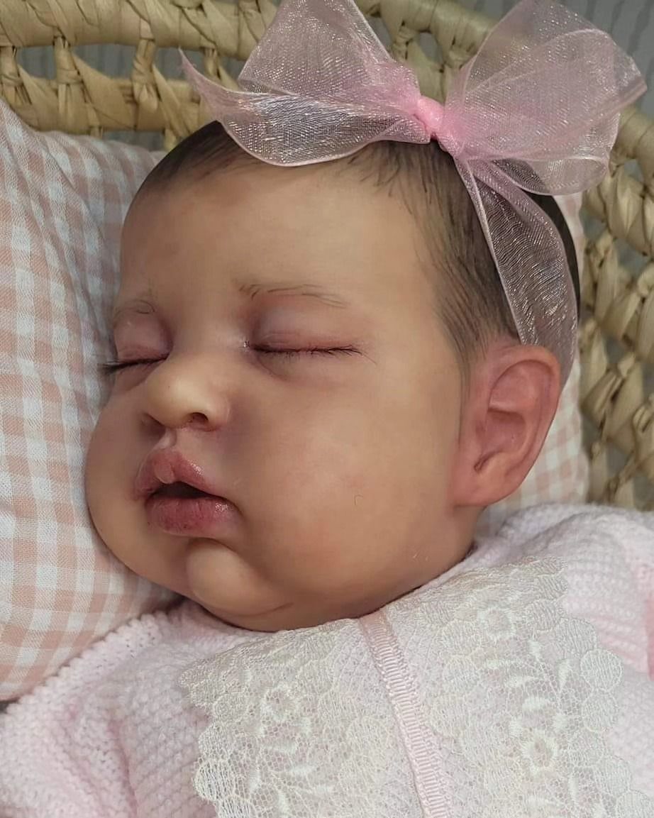Muñeca bebé durmiendo con un lazo rosa transparente y un conjunto de encaje blanco, descansando sobre un fondo tejido.