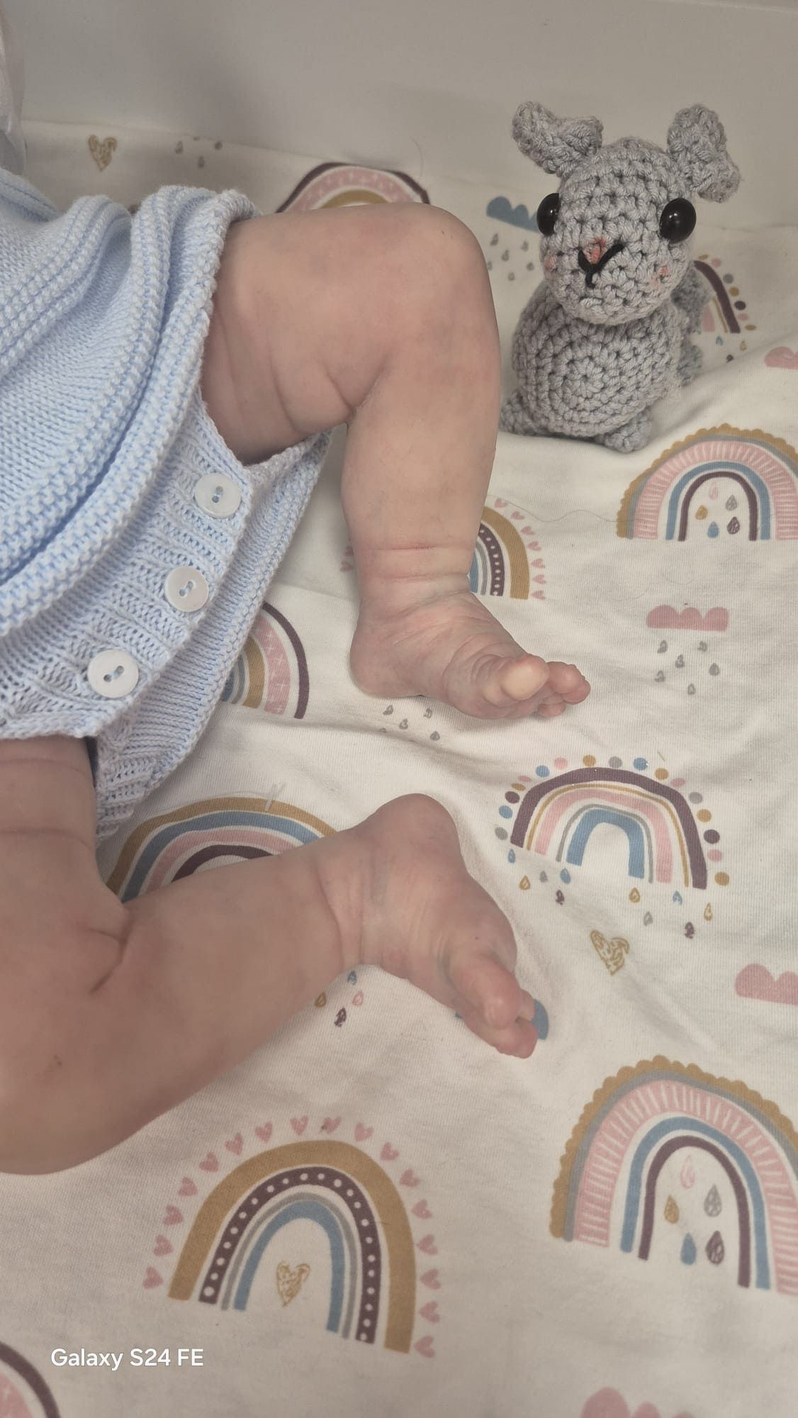 Un bebé con un conjunto azul sobre una manta con estampado de arcoíris, junto a un conejito de peluche gris tejido a crochet.