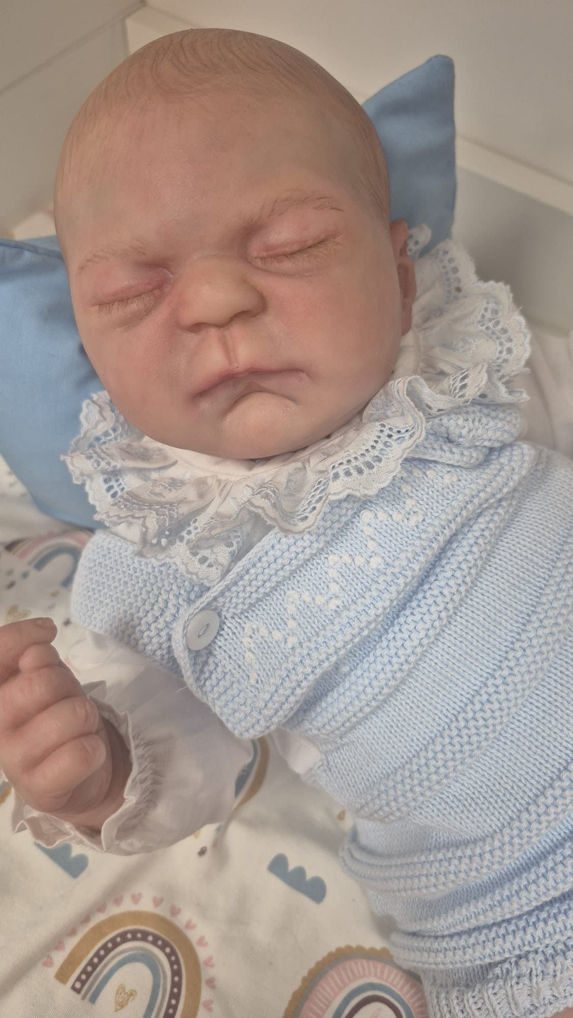 Muñeca bebé durmiendo, envuelta en una manta de punto azul, con un cuello de encaje y un chupete cerca.
