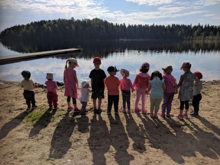 Värikkäisiin vaatteisiin pukeutuneet lapset seisovat järven rannalla ja katselevat tyynen veden yli metsäistä saarta.
