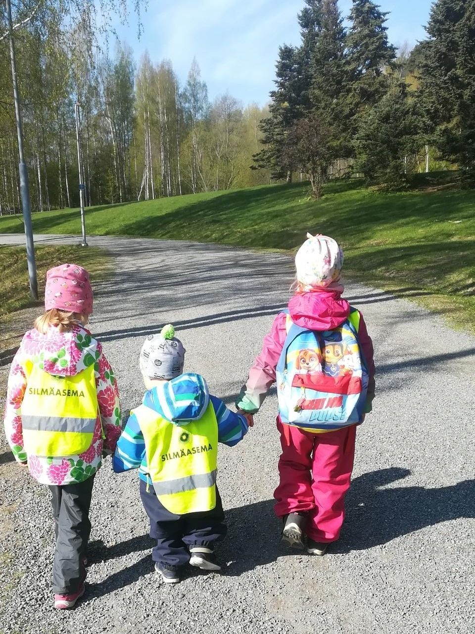 Kolme lasta kirkkaissa takeissa kävelee sorapolulla aurinkoisessa puistossa
