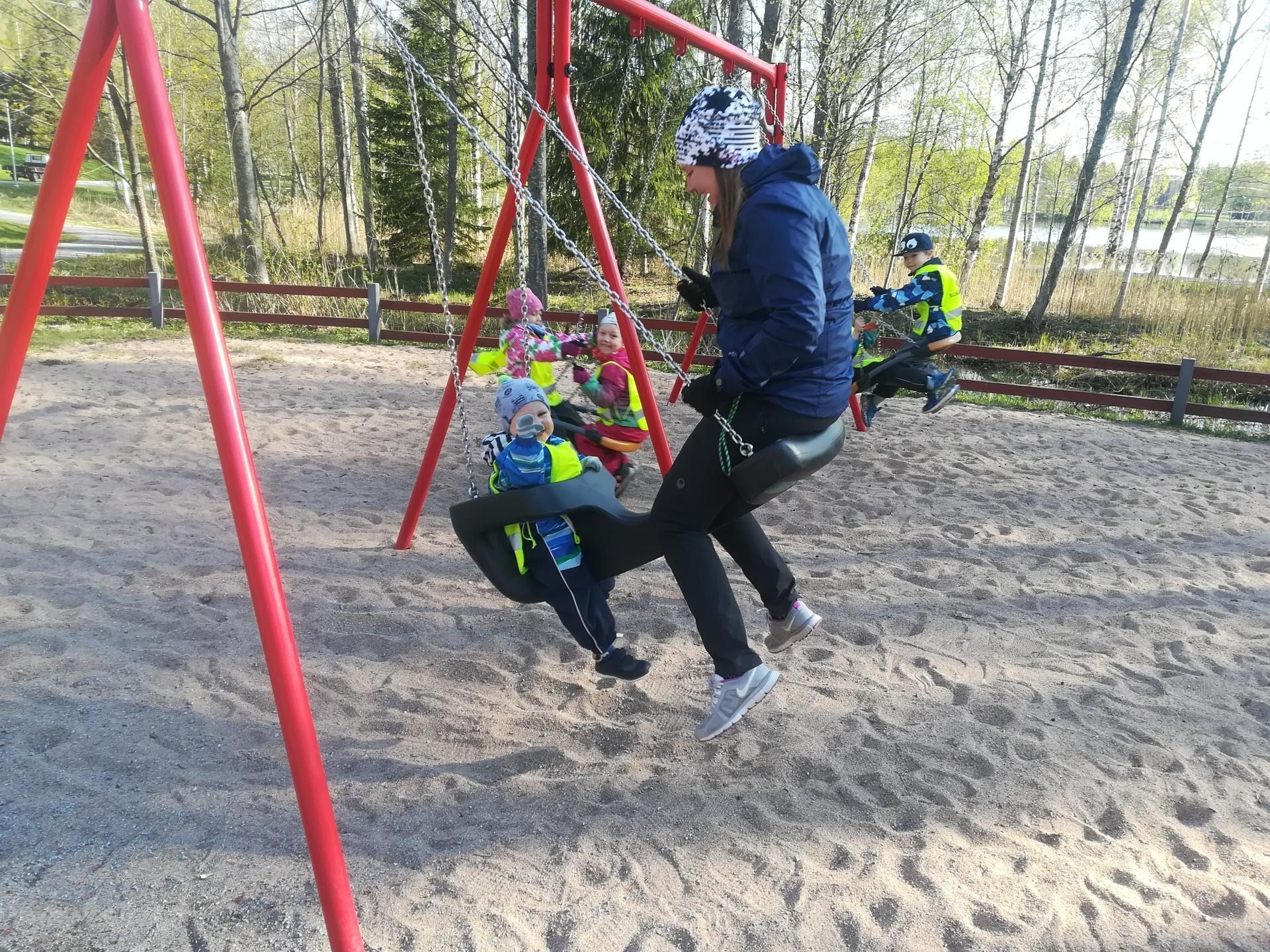 Lapsia leikkikentän keinussa, ja aikuinen työntää yhtä siniseen takkiin pukeutunutta lasta