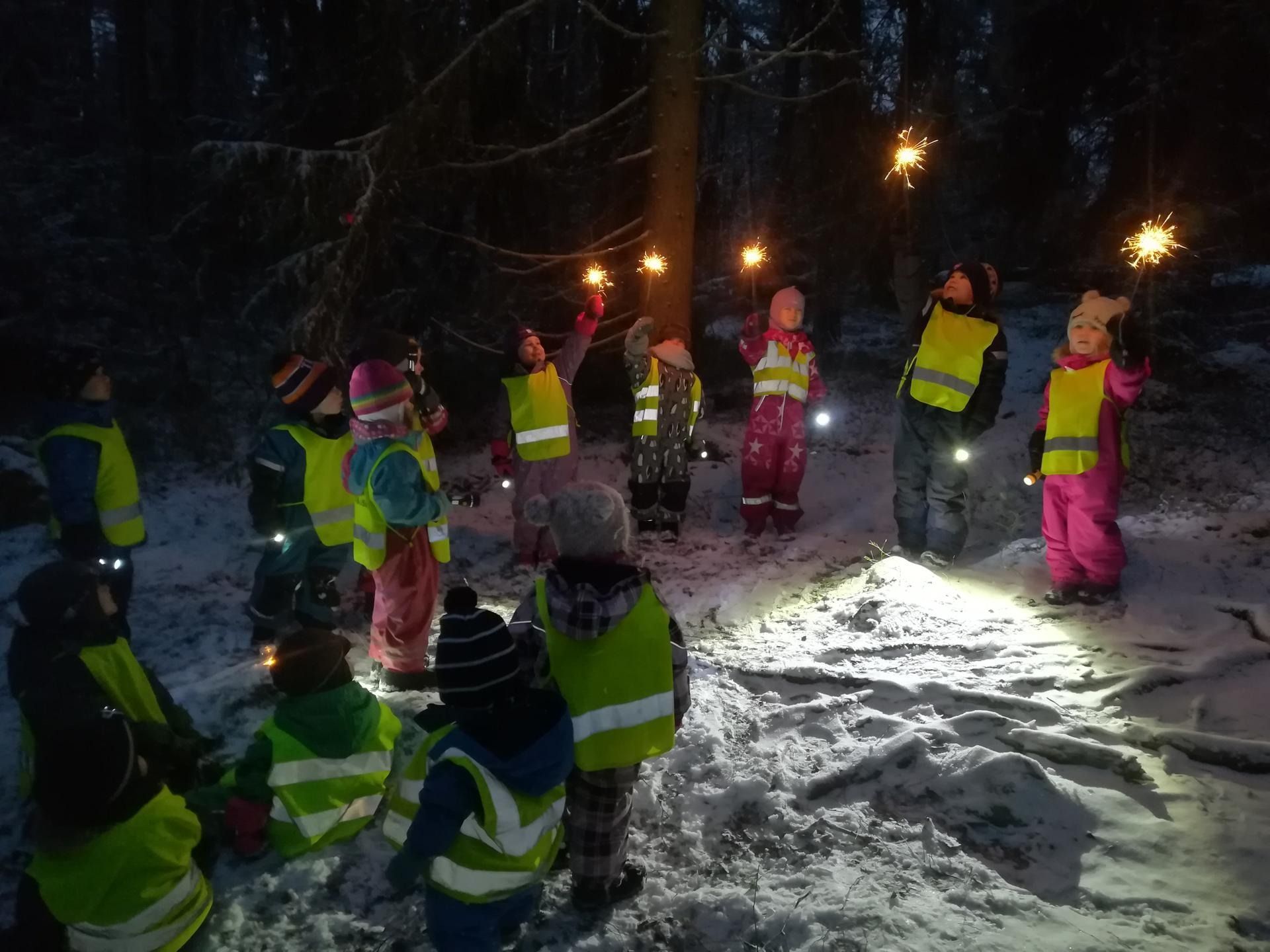 Kirkkaisiin talvivarusteisiin ja heijastinliiveihin pukeutuneet lapset kokoontuvat ulkona yöllä lumessa taskulamppujen kanssa