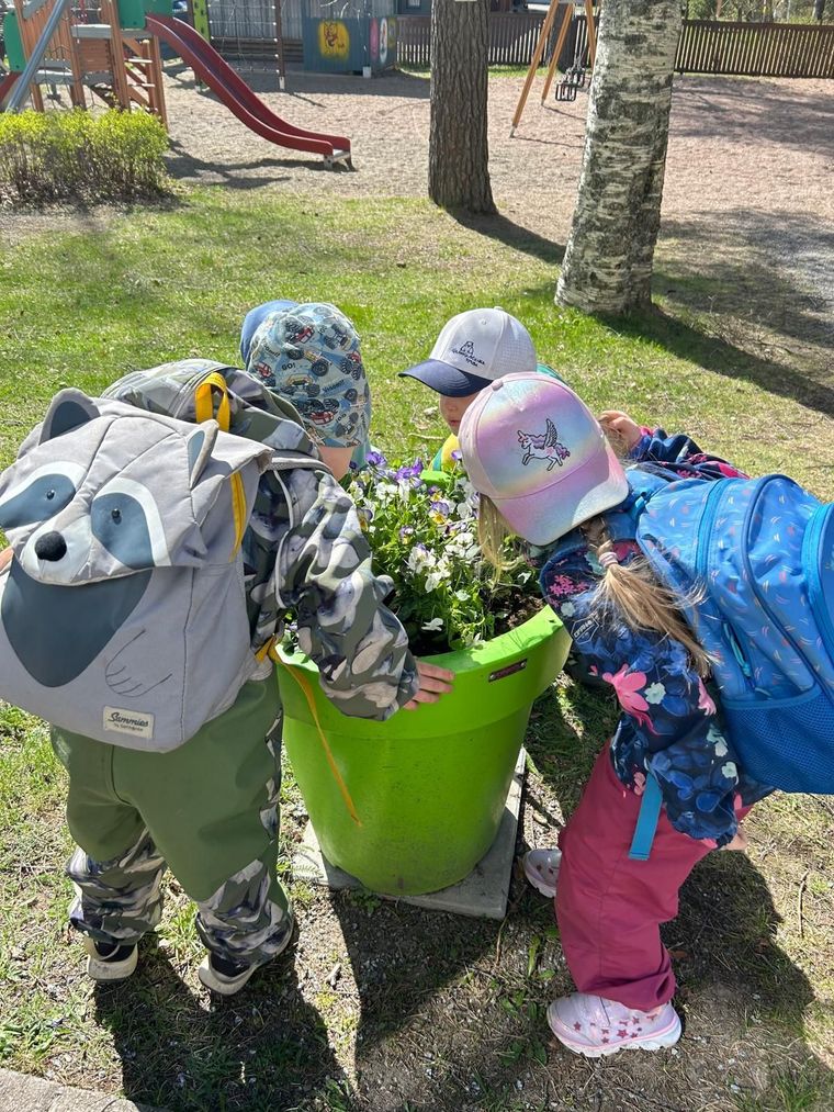 Värikkäisiin vaatteisiin pukeutuneet lapset nojaavat kirkkaanvihreän ruukun yli puistossa tai leikkikentällä.