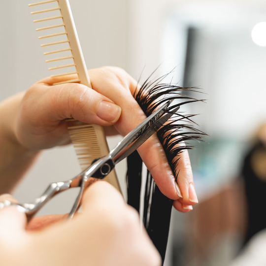 Mains coupant des cheveux foncés avec des ciseaux et un peigne dans un salon.