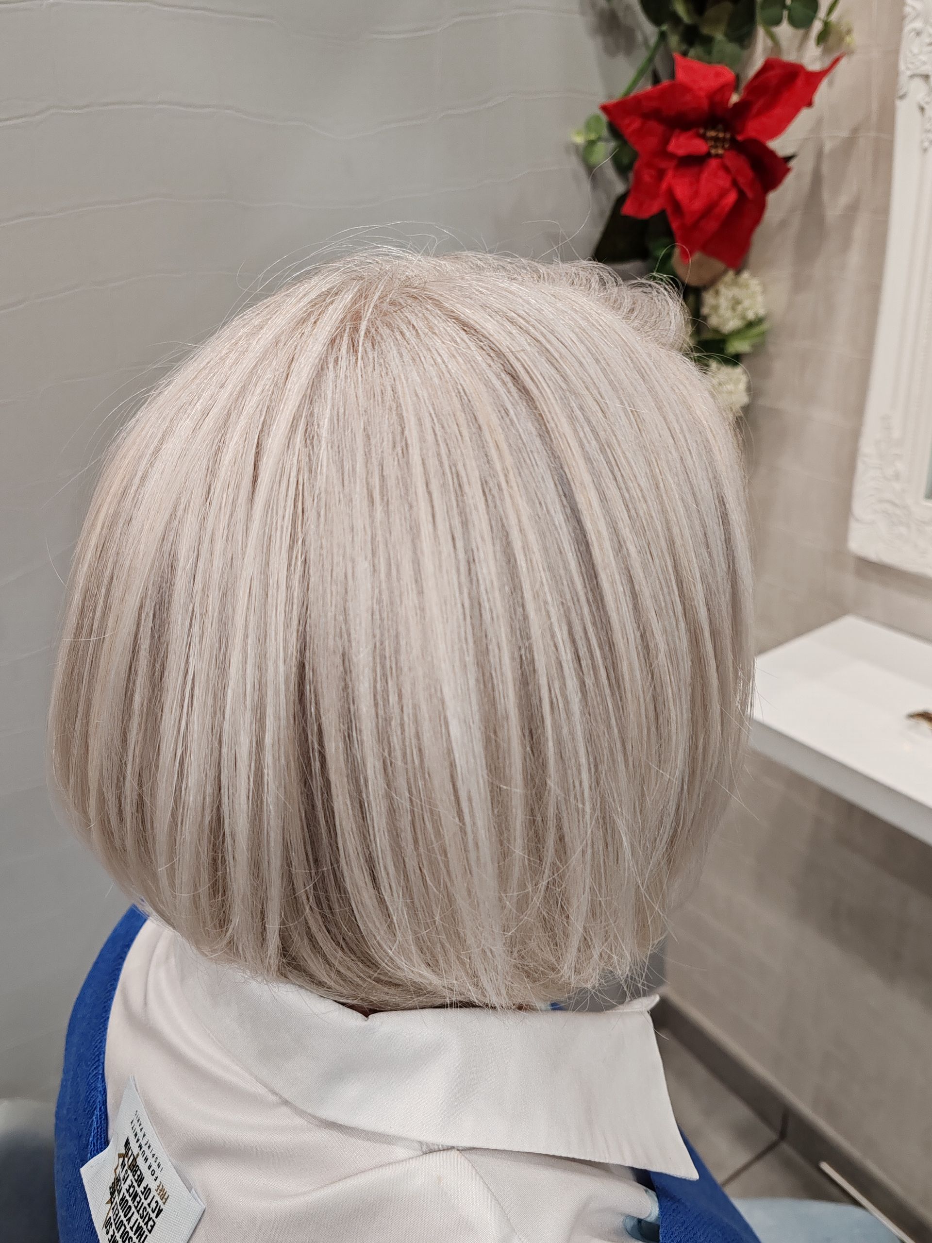 Coupe au carré, blonde vue de dos. Personne assise dans un fauteuil de salon, fleurs sur un mur en arrière-plan.