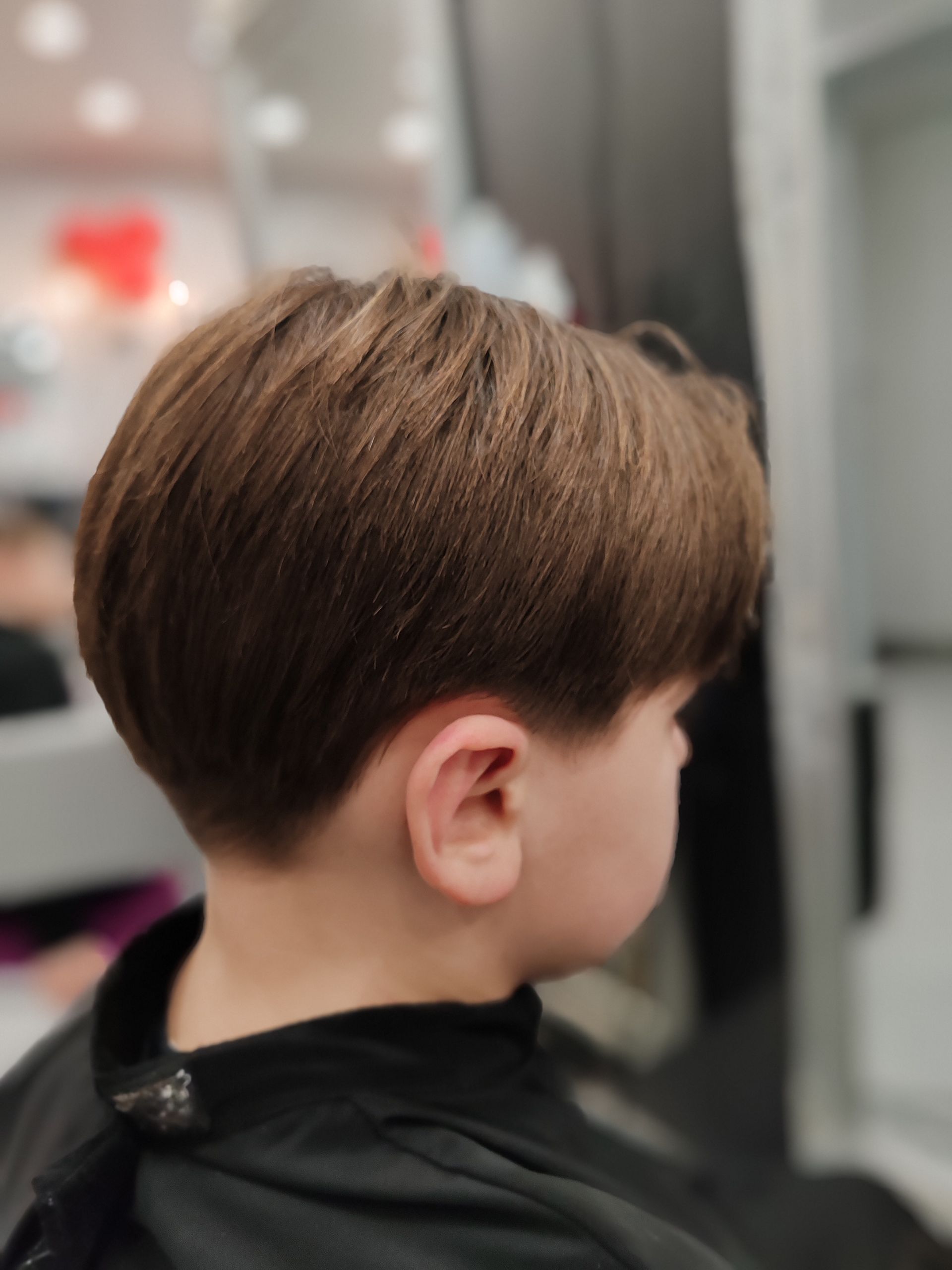 Garçon aux cheveux bruns, fraîchement coupés en forme de bol, dans un salon, portant une cape noire.