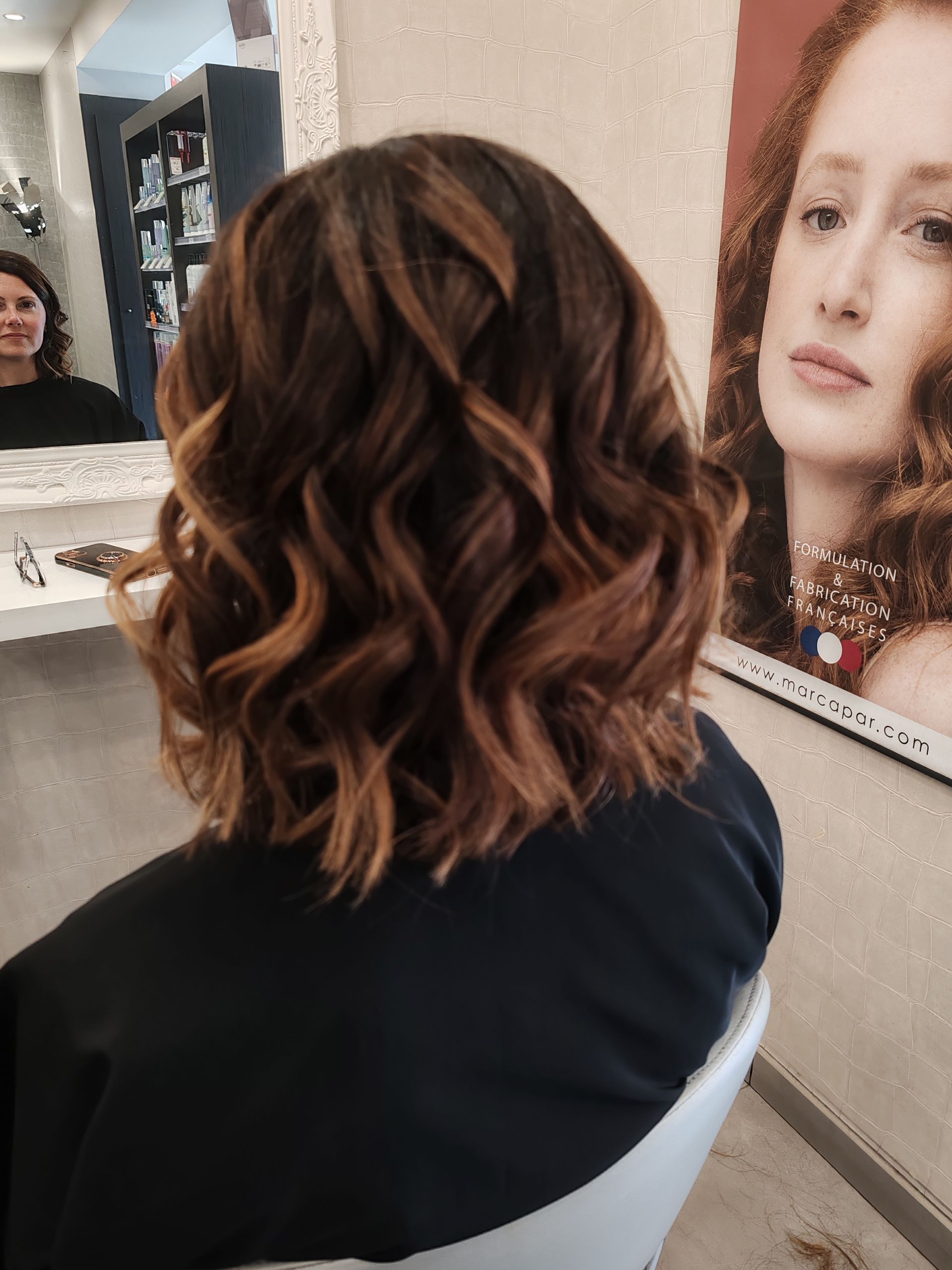 Femme aux cheveux bruns bouclés, coiffée au carré avec des mèches plus claires. Assise dans un salon de coiffure.