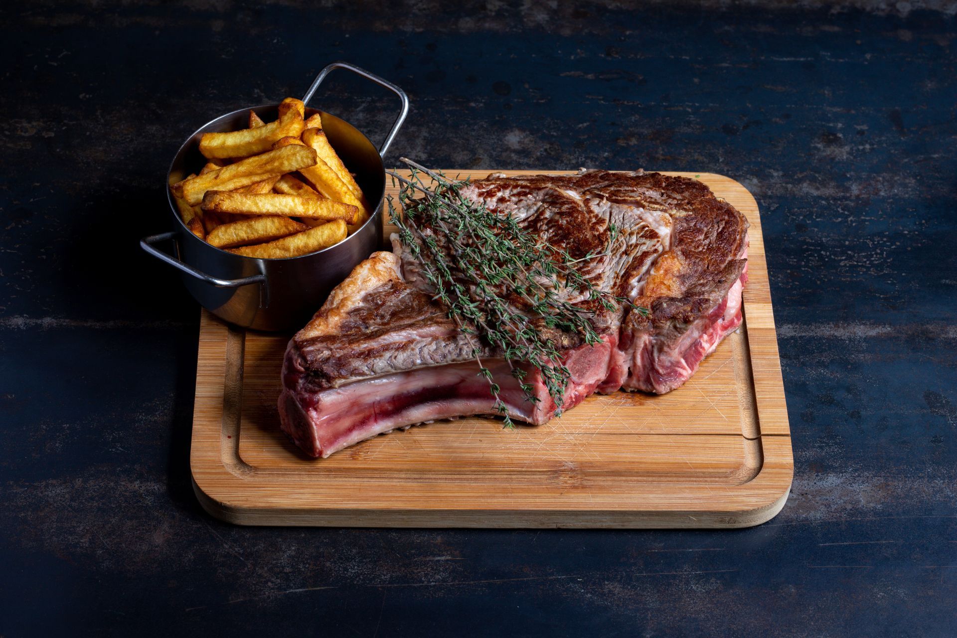 Une côte de boeuf sur une planche à découper avec des brins de romarin et une corbeille de frites sur le côté.