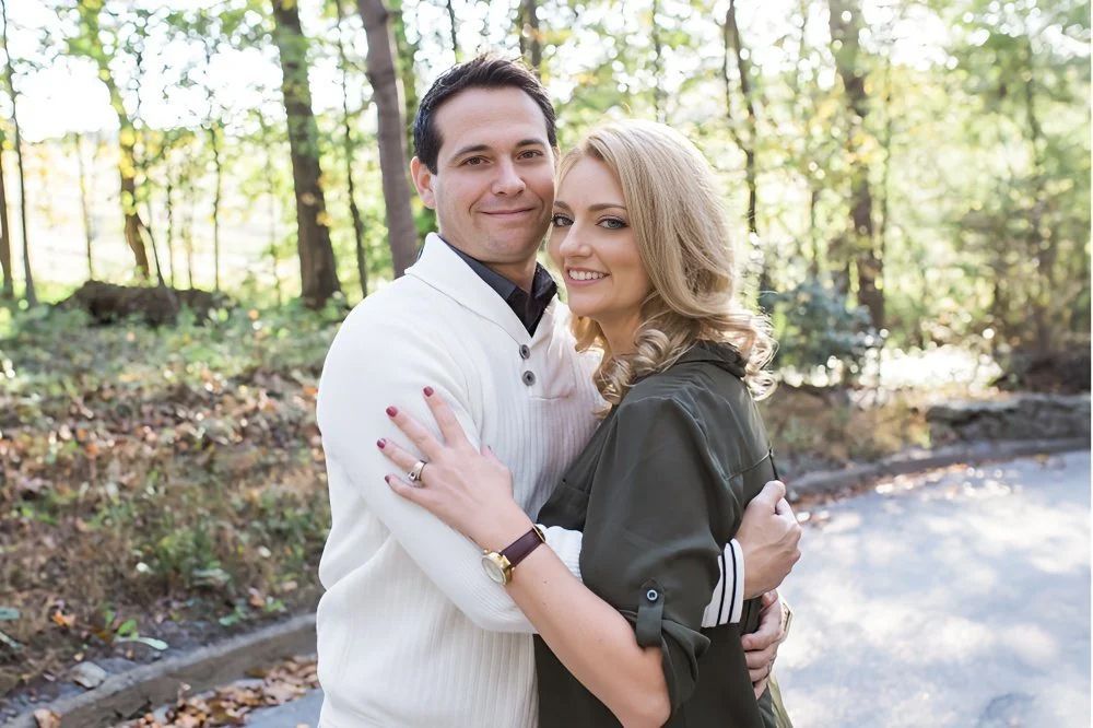 Couple hugging outdoors in front of trees. Man wearing white sweater, woman wearing olive green jacket.