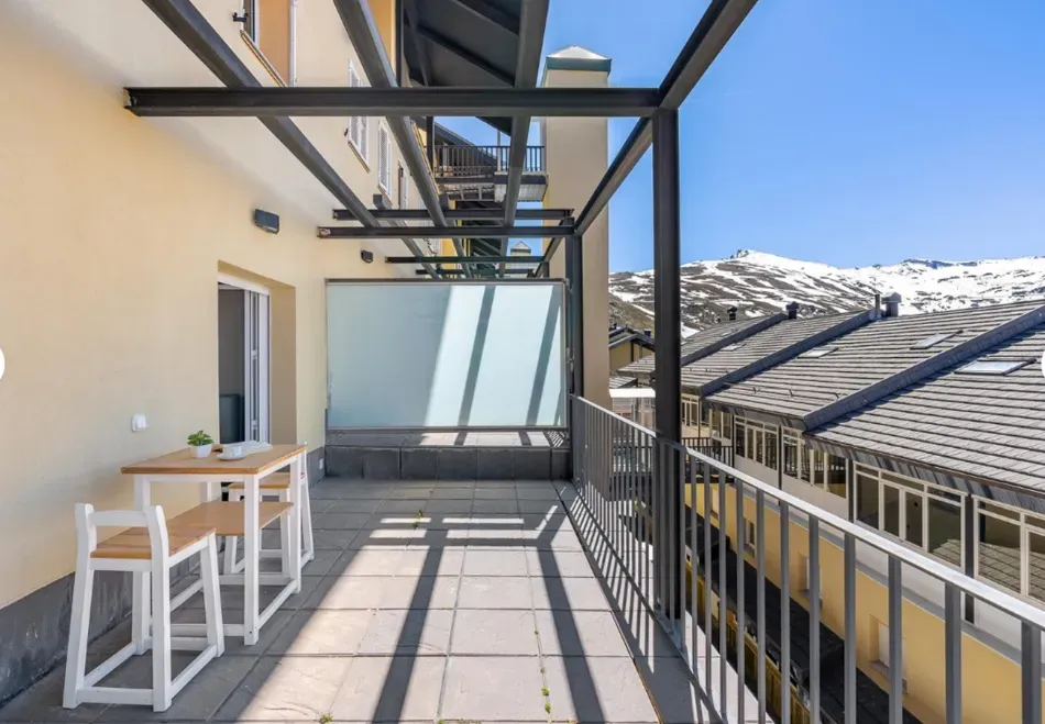 Terraza soleada en la azotea con mesa blanca, barandilla y vistas a la montaña más allá de los tejados vecinos.