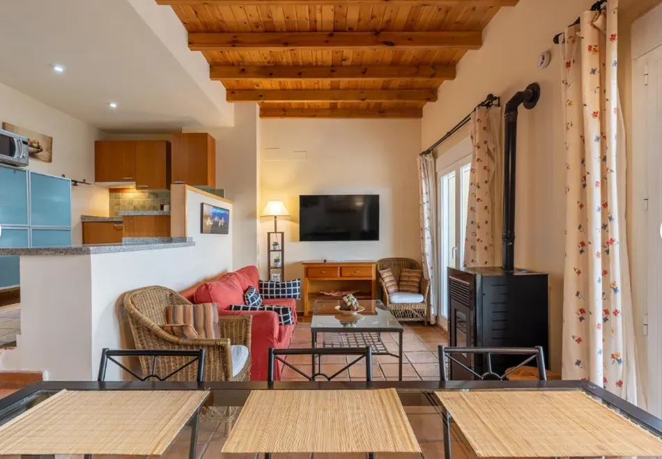 Acogedora sala de estar con techo de madera, televisor, chimenea, sillas de mimbre y cocina abierta al fondo.