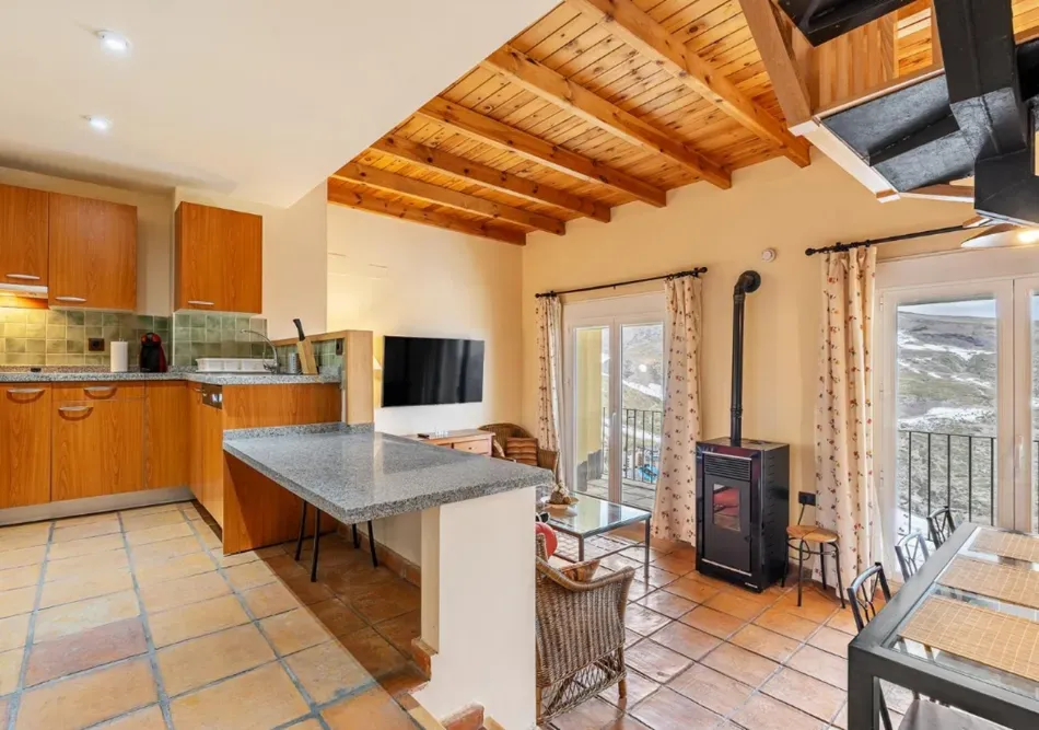 Cocina y comedor luminosos con vigas de madera, suelo de baldosas, isla de piedra y puertas de cristal que dan a un patio.