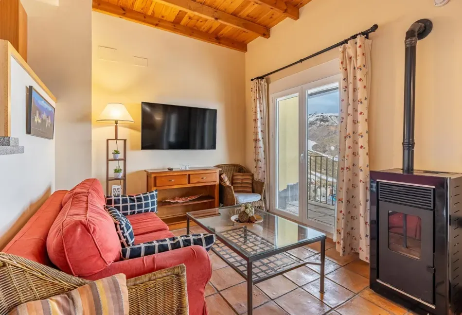 Acogedora sala de estar con sofá rojo, televisor montado en la pared, mesa de centro de cristal y estufa de leña junto a una puerta con ventana.