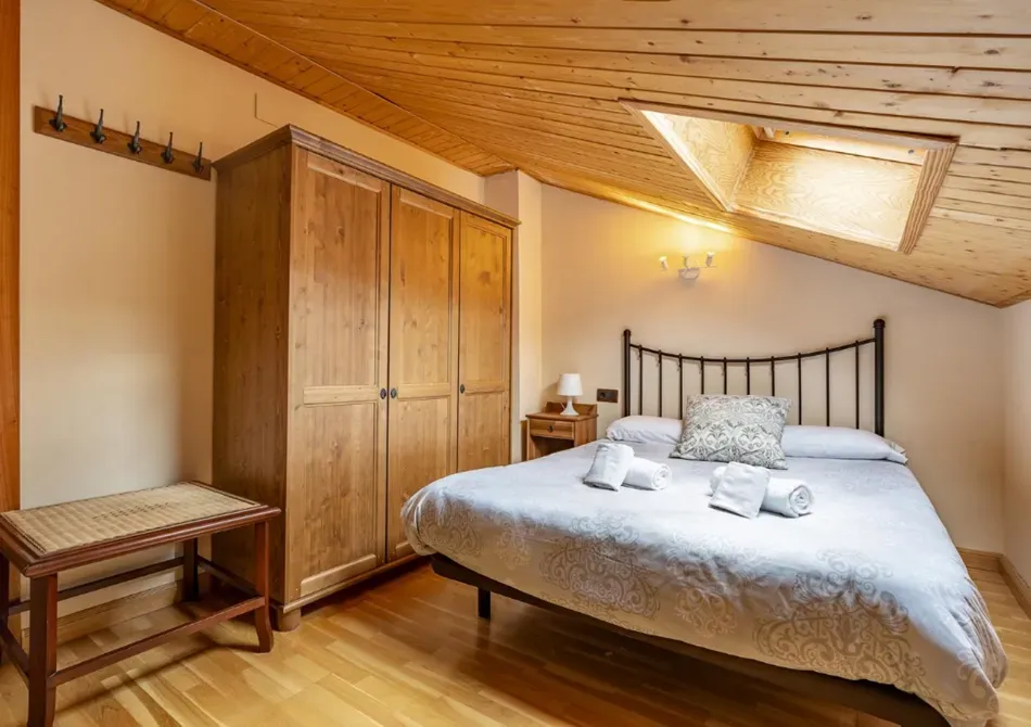 Acogedor dormitorio abuhardillado con techo de madera inclinado, armario de madera, banco y cama bien hecha.