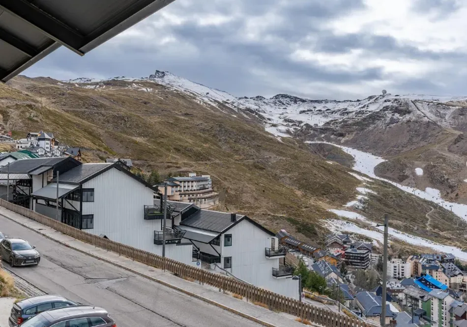 Pueblo de montaña nevado con casas blancas, una carretera y pistas de esquí bajo un cielo nublado.