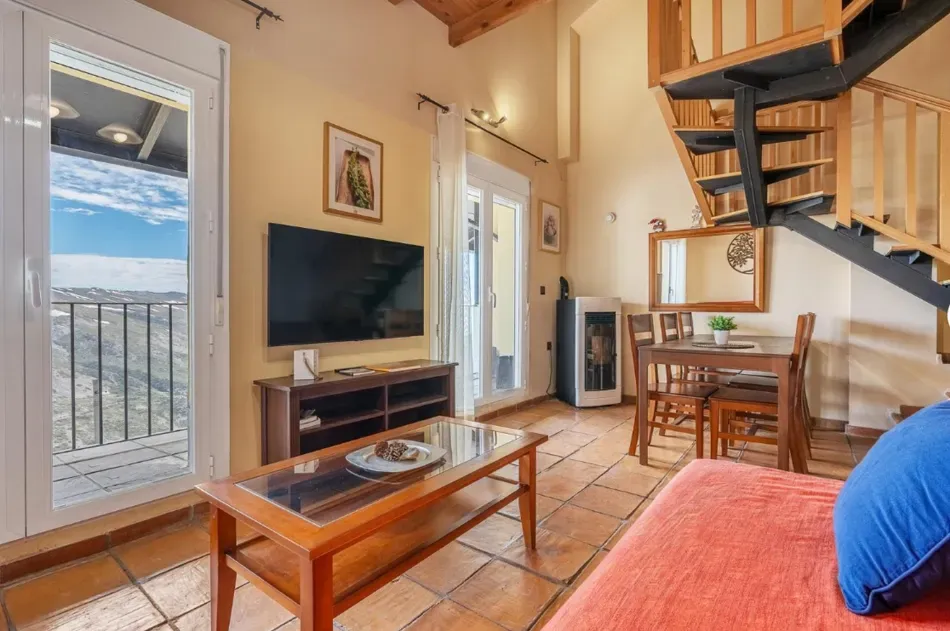 Acogedor salón tipo loft con sofá, televisor, mesa de centro, escalera de caracol y balcón con vistas.