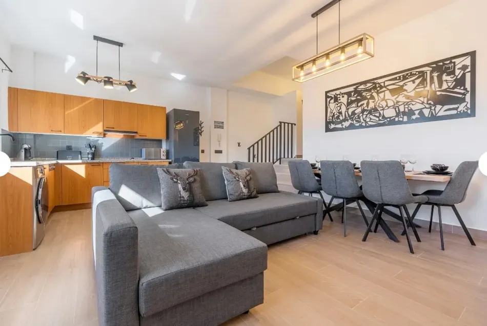 Sala de estar de planta abierta con sofá seccional gris, mesa de comedor y cocina con gabinetes de madera.