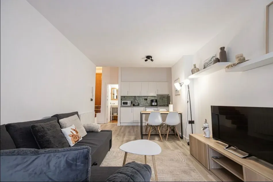 Sala de estar moderna con sofá gris, televisor y un pequeño comedor junto a una cocina luminosa.