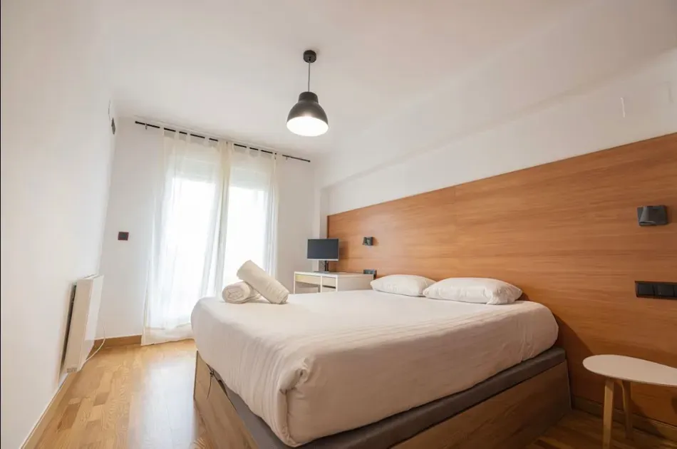 Dormitorio luminoso y minimalista con cama king size, pared decorativa de madera y cortinas que dejan entrar la luz natural.