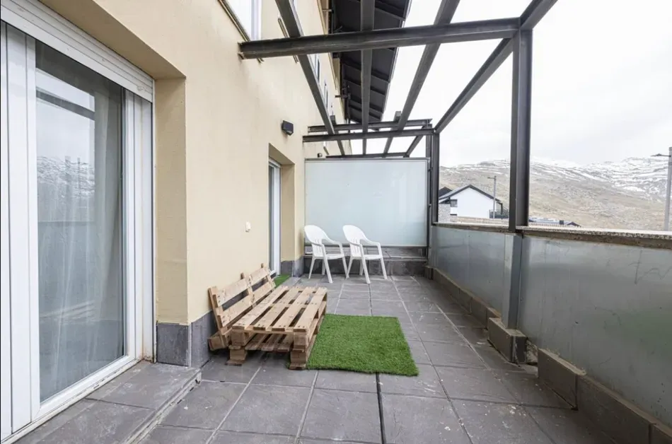 Pequeño balcón con suelo de baldosas grises, barandilla de cristal, sillas de patio y palets de madera apilados bajo una pérgola.