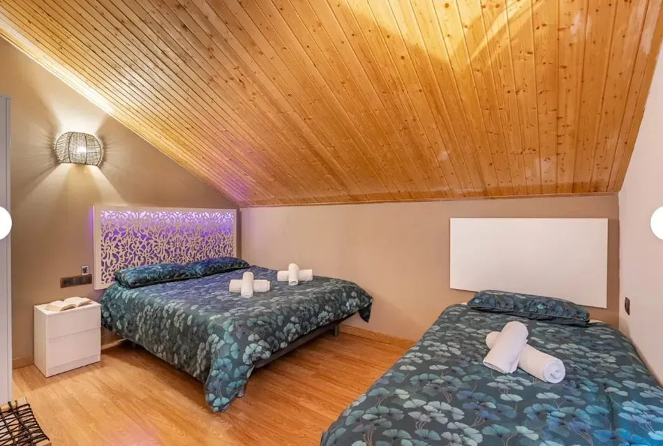 Habitación tipo loft con dos camas, techo abovedado revestido de madera e iluminación cálida en las paredes.
