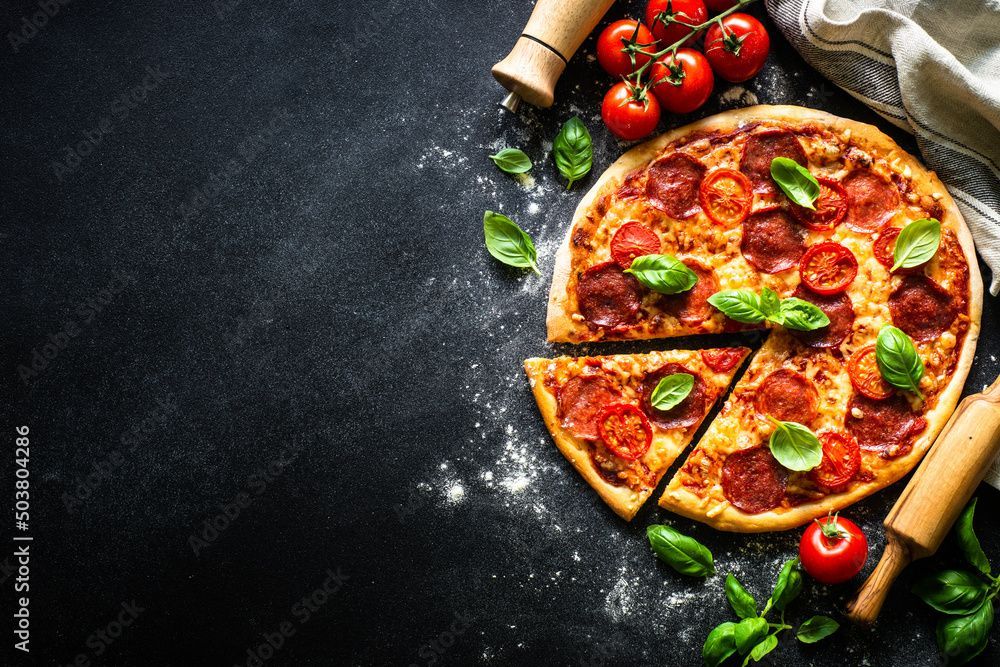 Pizza con peperoni, pomodori e basilico su una superficie scura, parzialmente tagliata.