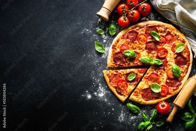 Pizza con peperoni, pomodori e basilico su una superficie scura, parzialmente tagliata.