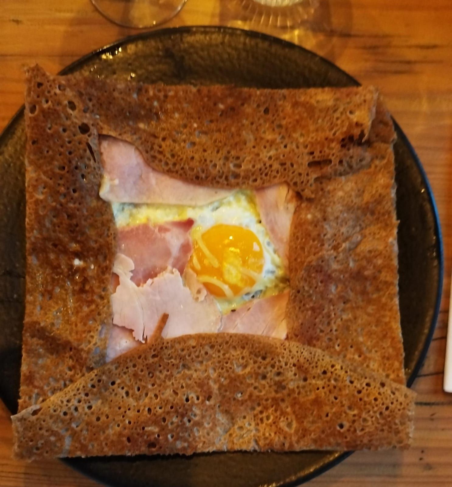Galette salée au jambon, œuf et jaune doré. Crêpe brune et croustillante, pliée en carré sur une assiette noire.