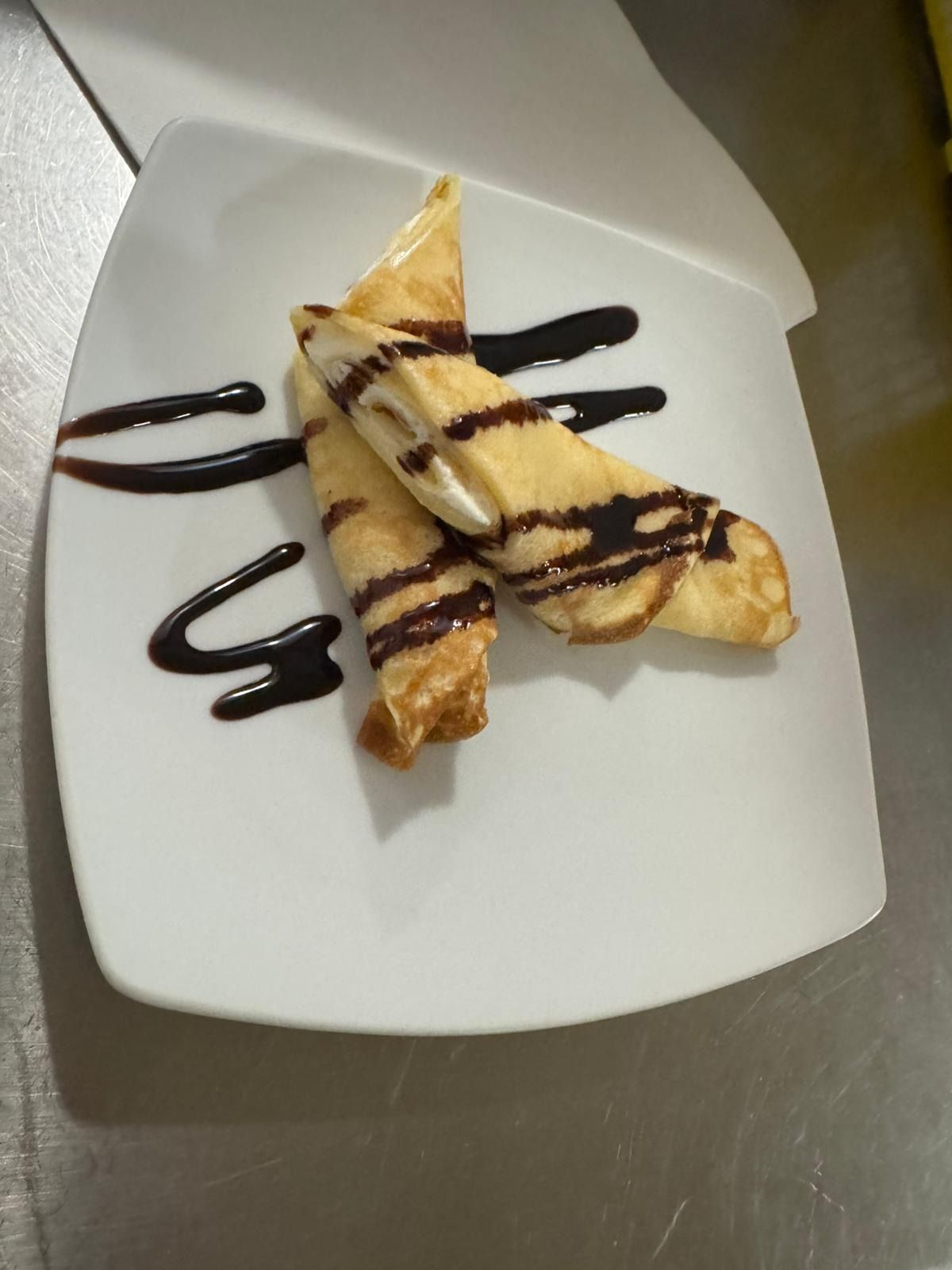 Dos crepes enrollados rellenos de crema blanca, rociados con salsa de chocolate, servidos en un plato blanco.