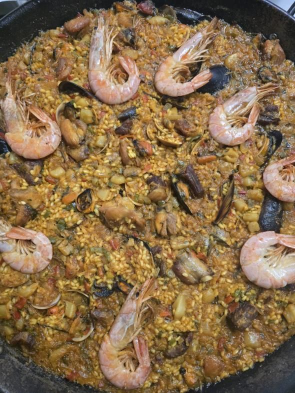 Una paella grande cubierta con gambas cocidas, mejillones y trozos de carne, servida en una sartén de hierro fundido oscuro.