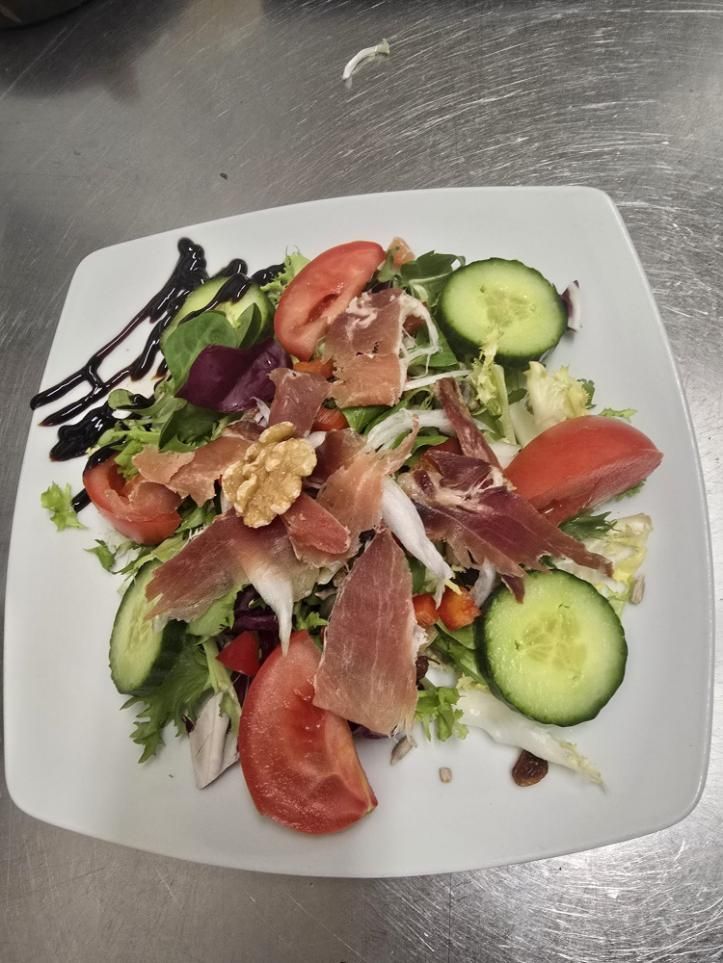Una ensalada servida en un plato cuadrado blanco con rodajas de tomate, pepino, jamón curado y nueces, aderezada.