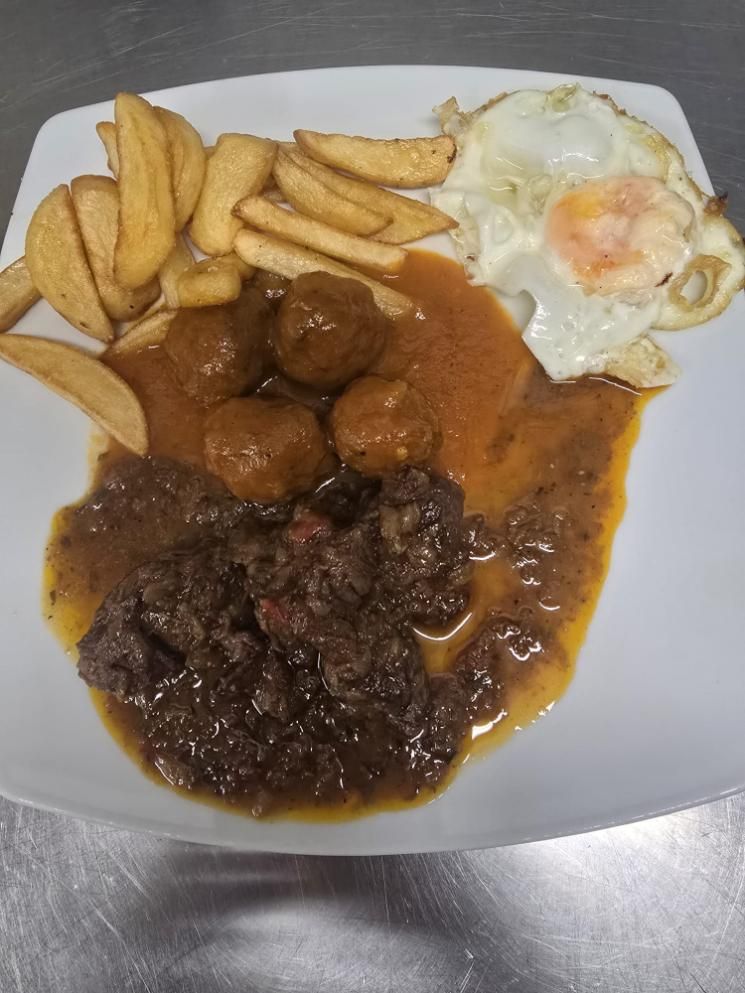 Un plato blanco con albóndigas en salsa, un estofado de ternera, patatas fritas en gajos y un huevo frito
