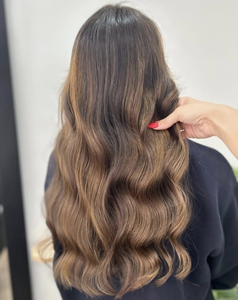 Mujer con cabello castaño largo y ondulado, vista de espaldas. Lleva un top oscuro; al fondo, un estante blanco y una planta.
