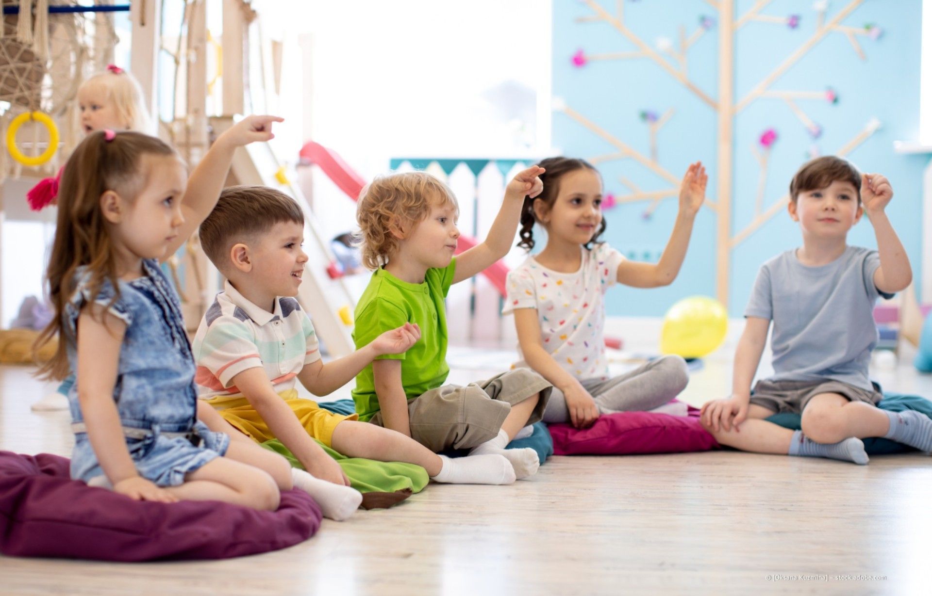 Kinder sitzen in einem hellen Klassenzimmer auf dem Boden und heben während einer Gruppenaktivität die Hände.