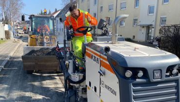 Ein Straßenarbeiter in orangefarbener Schutzkleidung asphaltiert eine Straße neben einer Straßenwalze in einem Wohngebiet.