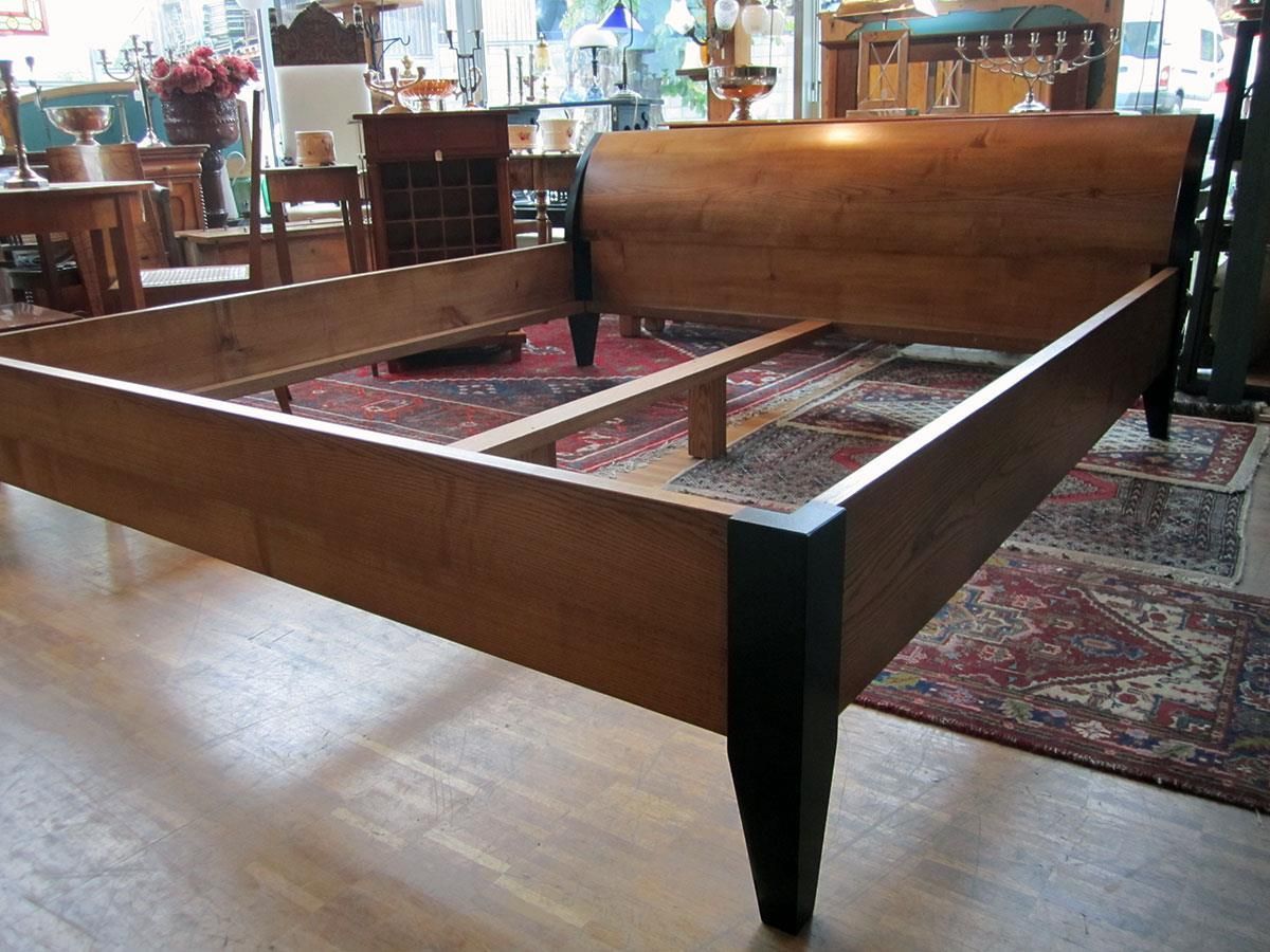 Ein hölzerner Bettrahmen mit schwarzen Beinen steht auf einem Holzboden