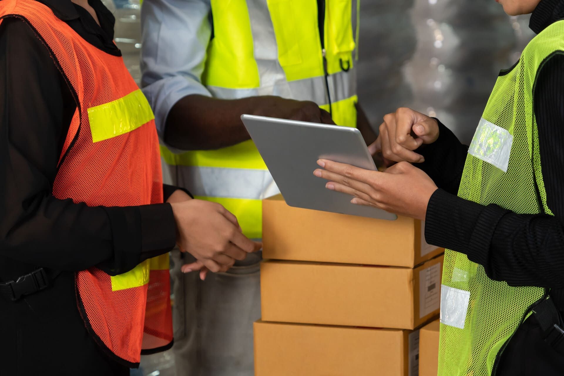 Trabajadores de almacén con chalecos de seguridad usando una tableta junto a cajas de cartón apiladas