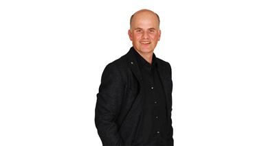 Ein kahlköpfiger Mann in einer schwarzen Jacke und einem schwarzen Hemd steht vor einem weißen Hintergrund.