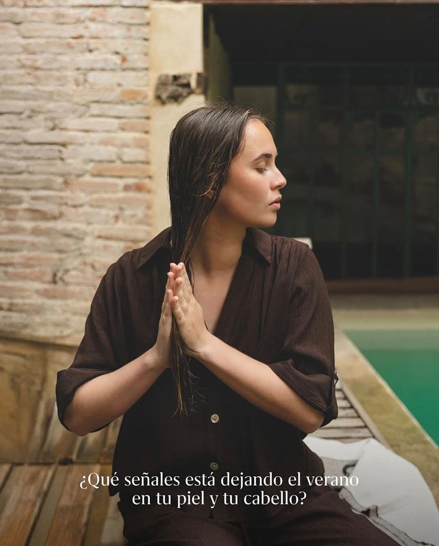 Mujer con el pelo trenzado, vestida con una túnica marrón, con las manos juntas junto a un estanque en un patio de piedra.