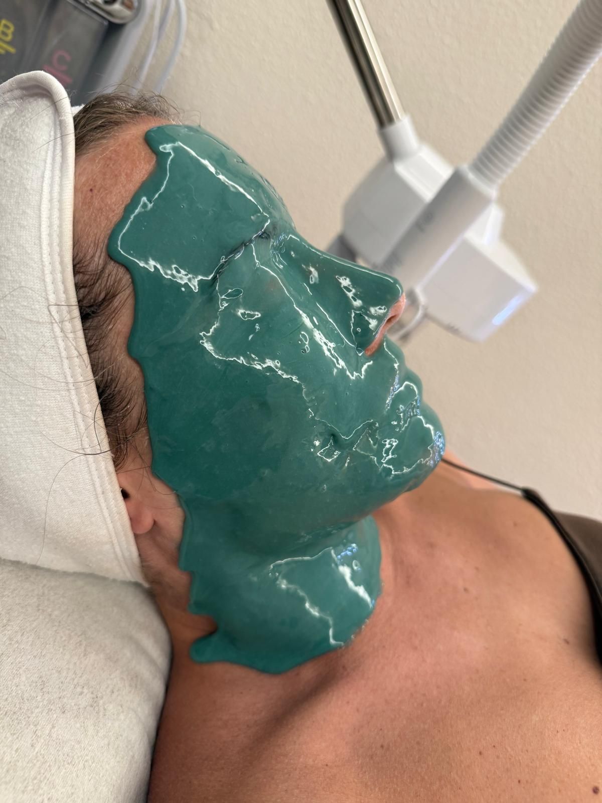 Persona recibiendo un tratamiento con mascarilla facial verde bajo el brazo de una máquina de spa