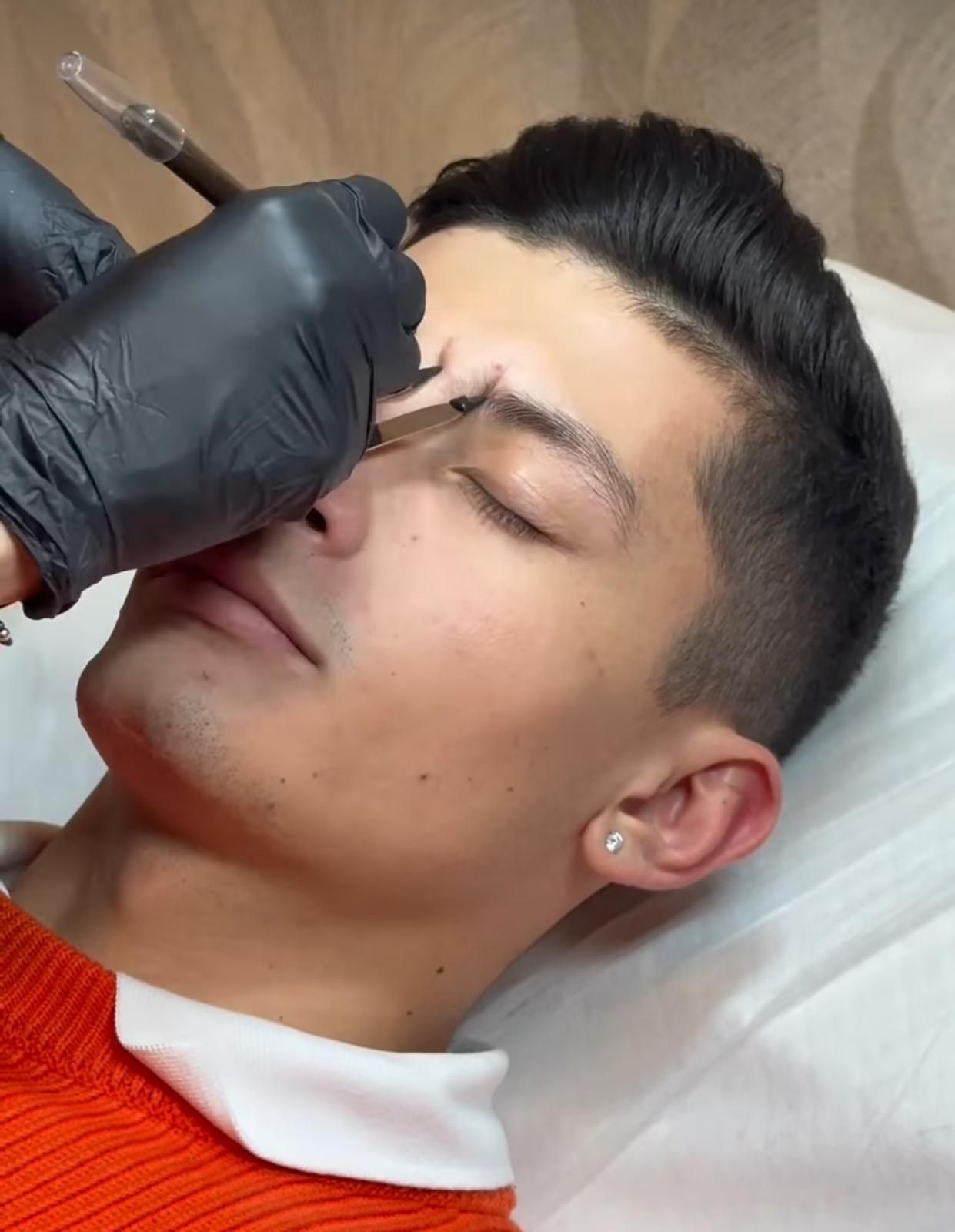Una mano enguantada dibuja una línea en la ceja de un hombre con un bolígrafo de tatuaje durante un procedimiento cosmético.