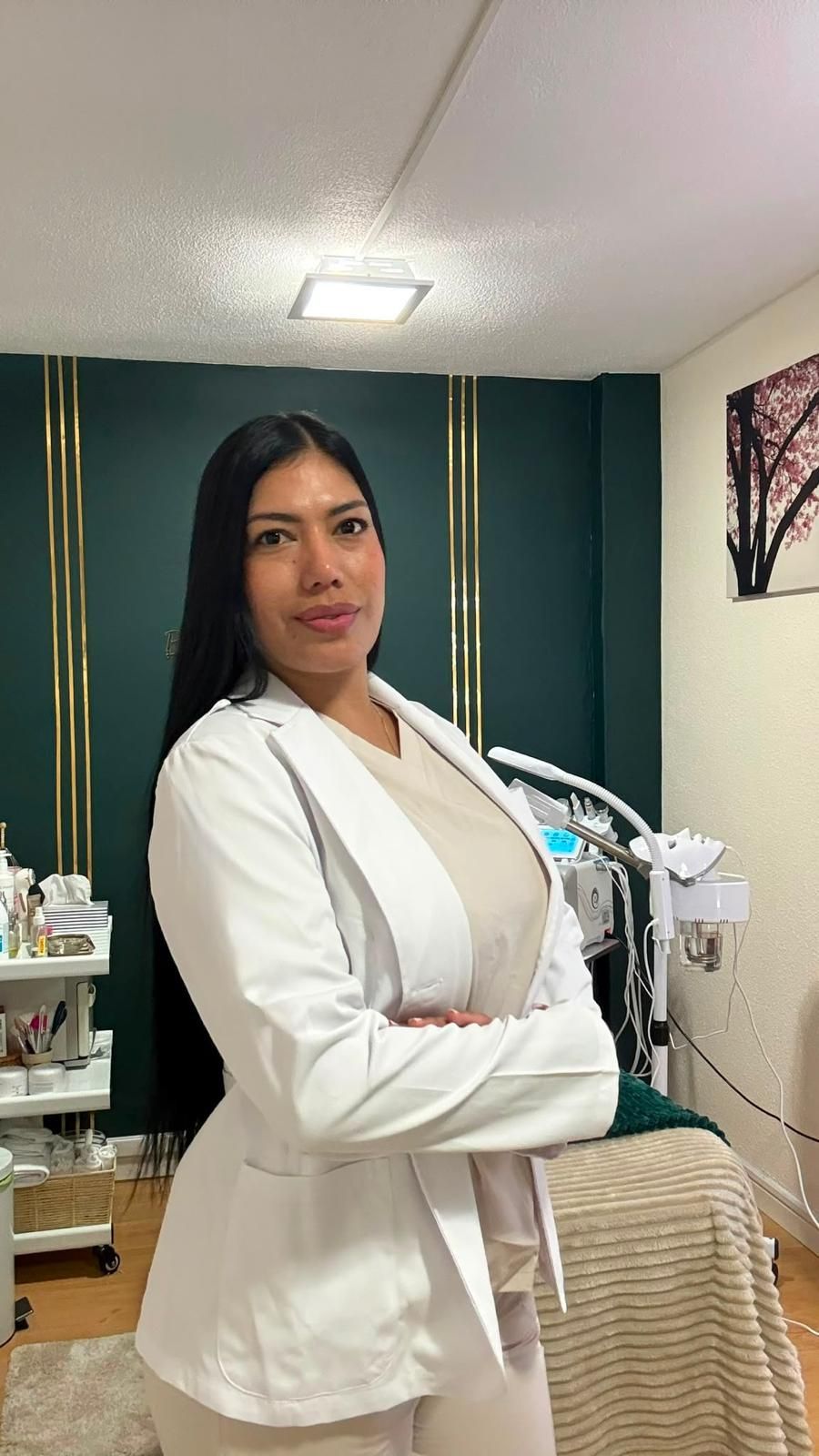 Mujer con chaqueta blanca de pie en una sala de spa o clínica, junto a un equipo de tratamiento, mirando a la cámara.