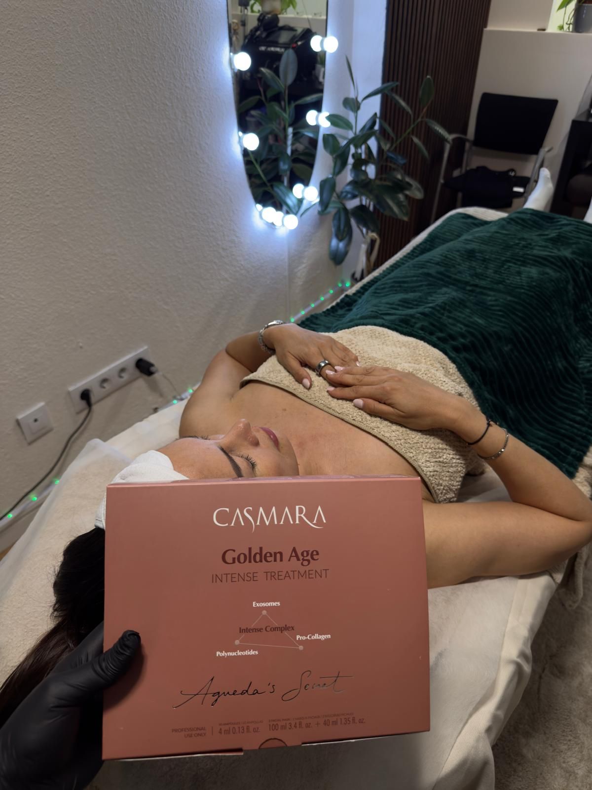 Una persona tumbada en una camilla de tratamiento con una mascarilla facial, sosteniendo en primer plano una caja rosa de productos para el cuidado de la piel.