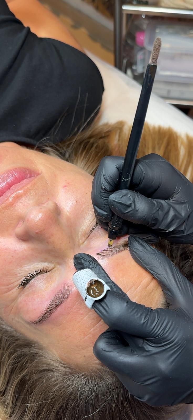 Primer plano de una persona a la que le están aplicando maquillaje de cejas con un pincel fino mientras está tumbada.